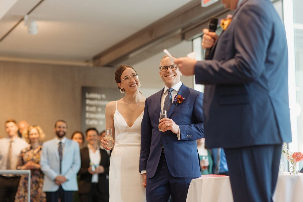 Bride and groom laughing while standing next to each other as best man gives speech, at their Saint Louis Contemporary Art Museum wedding.