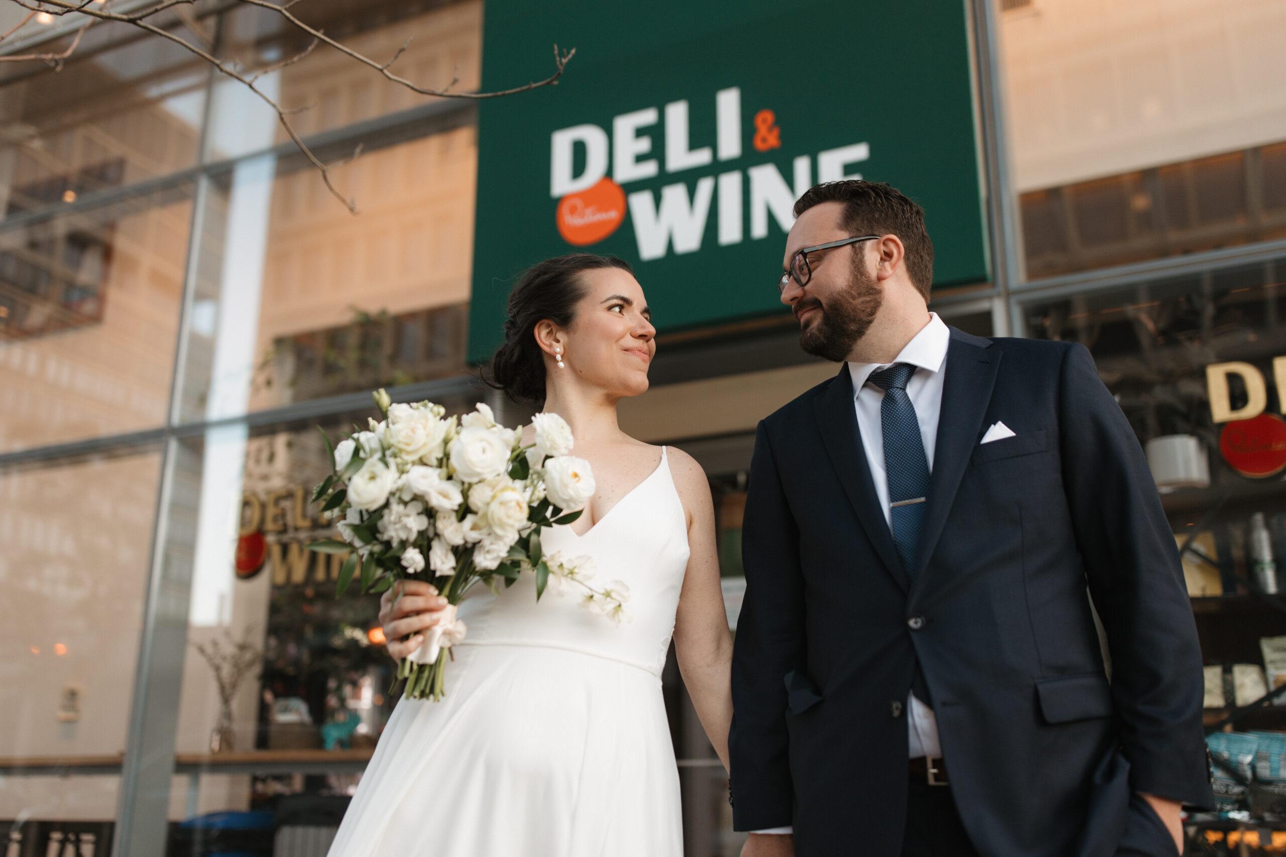 Bride with flowers and groom stand in front of Pastaria Deli & Wine wedding