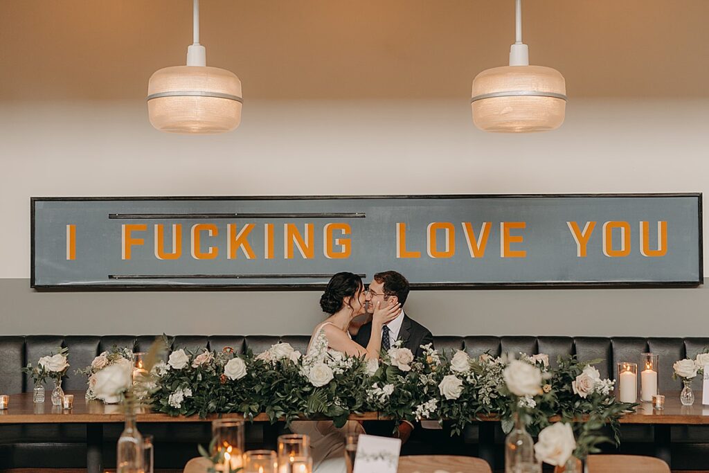 A bride and groom kiss while sitting under a sign that says "I fucking love you."