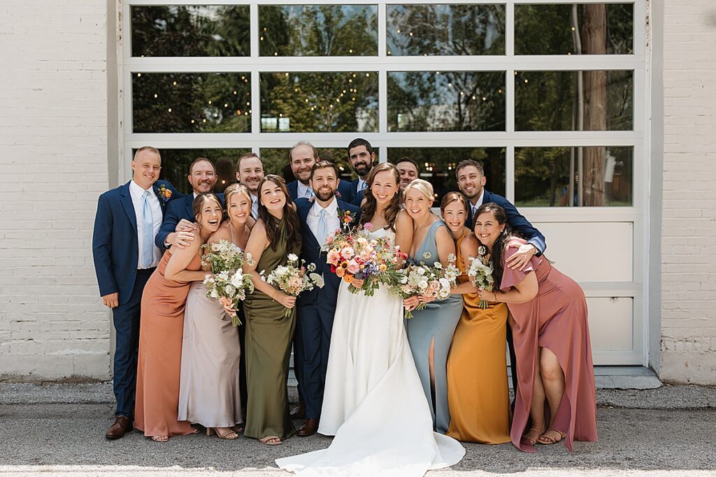 A wedding party wearing colorful dresses and navy suits smiling in front of a garage door.