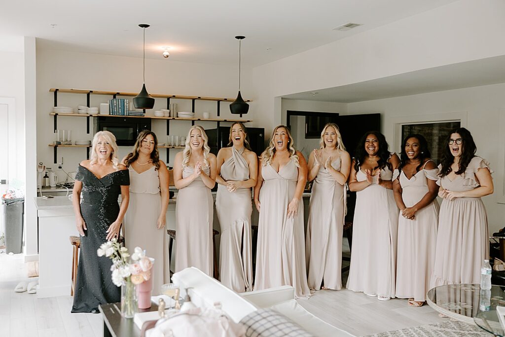 A group of bridesmaids wearing blush dresses looking surprised to see the bride in her dress.