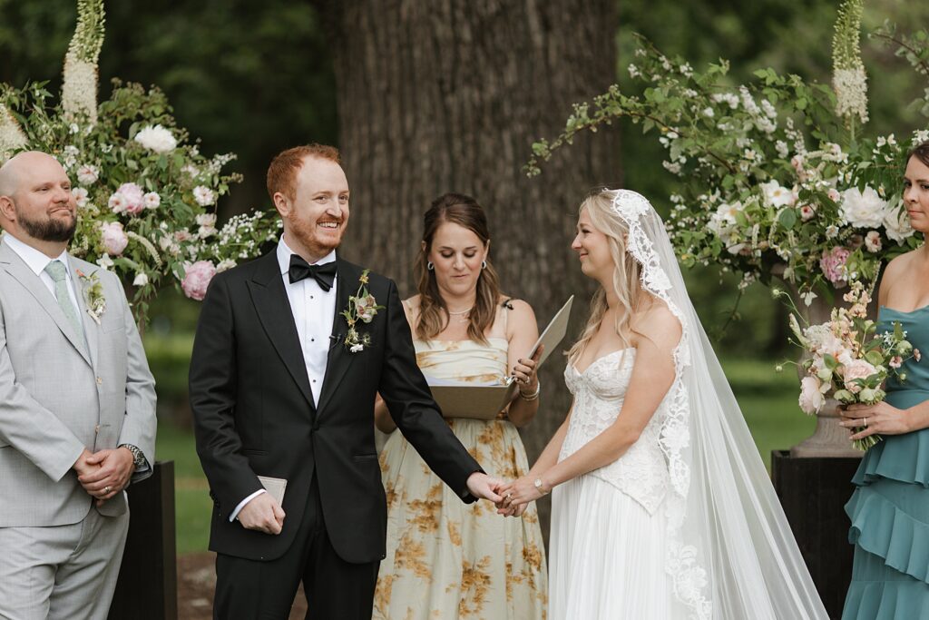 Bride and groom look out at crowd of guests, in front of large floral display, during ceremony at Weddings on Magnolia in St. Louis, Missouri